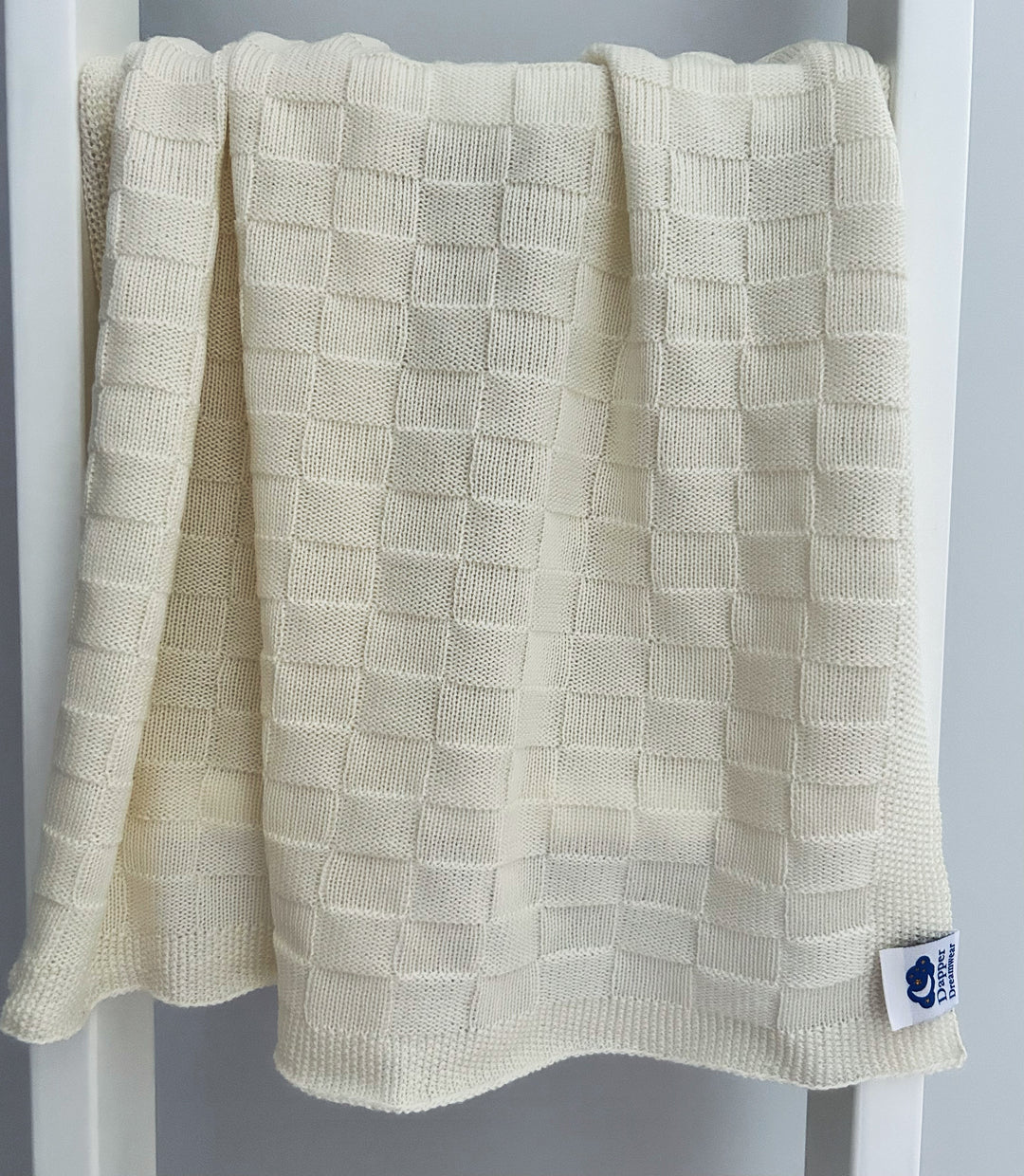 A white, solid-colored merino wool baby blanket with a textured design, draped over the back of a chair. Made in Australia.