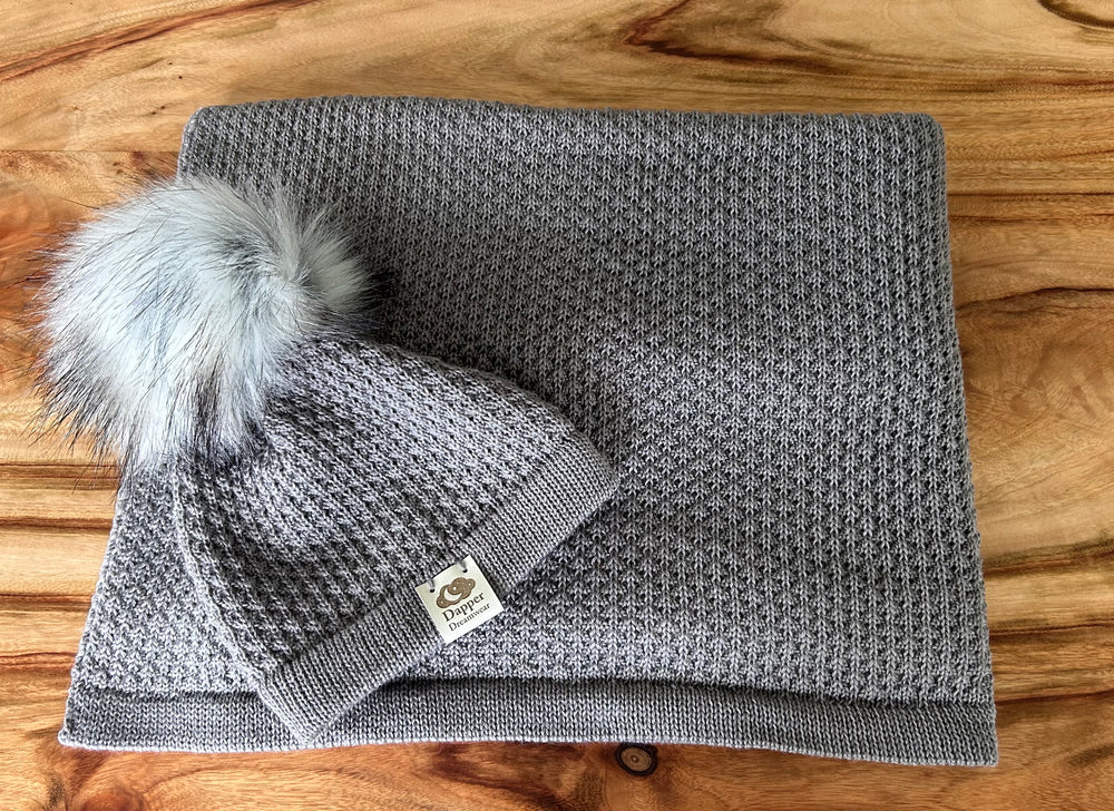 A grey merino wool baby blanket with a textured pattern and a matching beanie with a faux fur pom pom.