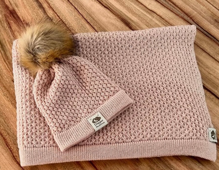 A honey coloured merino wool baby blanket with a textured pattern and a beanie with a pom-pom, displayed on a wooden surface.